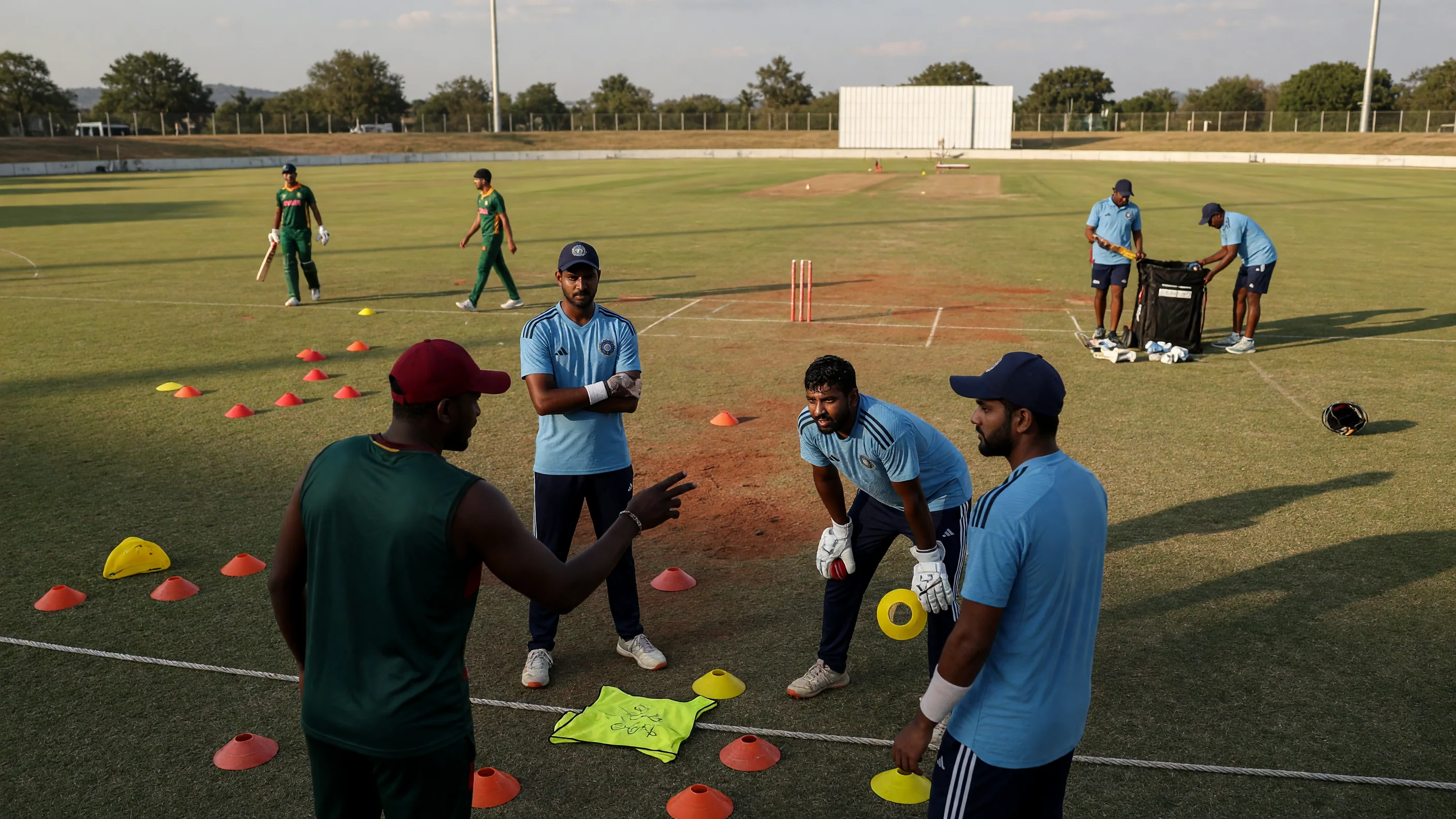 Fielders set in a ring on a sunlit outfield with cones and practice gear near the square